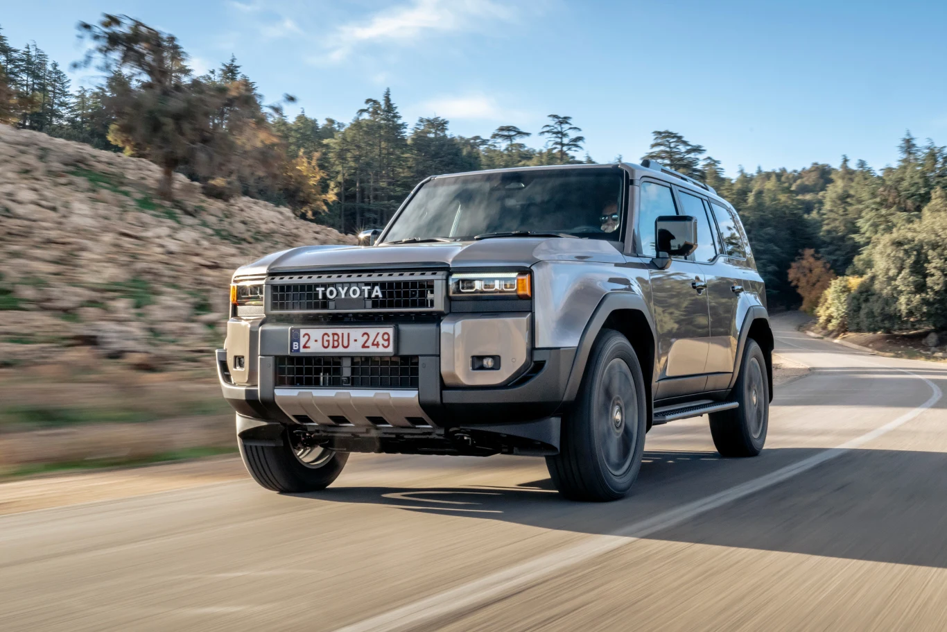 Toyota Land Cruiser w wersji Executive. Szara Toyota Land Cruiser w ruchu na krętej, asfaltowej drodze, otoczona kamienistym terenem i lasem w tle.