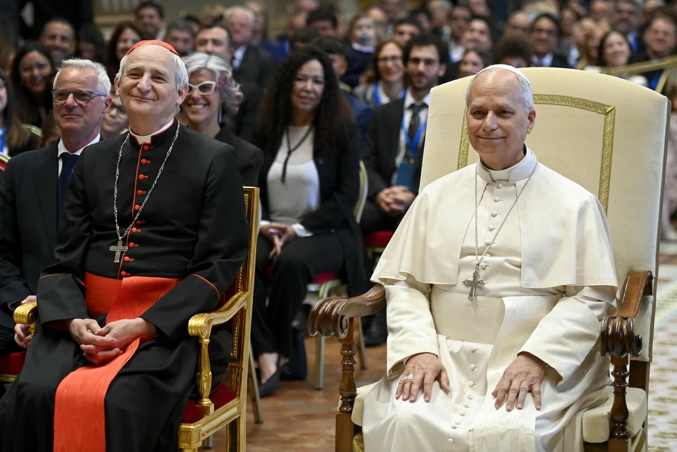Dwóch dostojników kościelnych w szatach liturgicznych siedzących na ozdobnych krzesłach na tle uśmiechniętej publiczności.