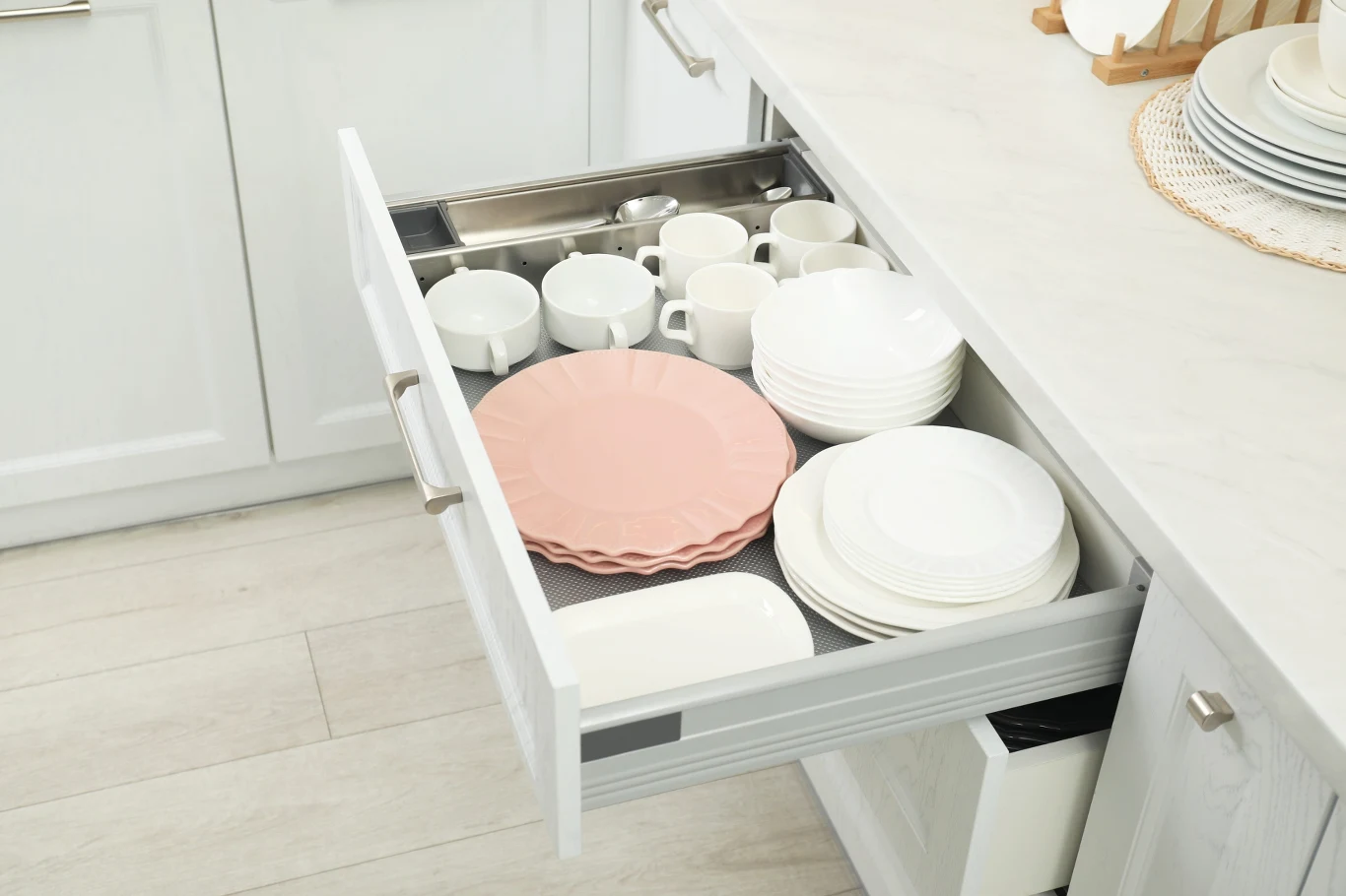Uchylona szuflada w białej kuchennej zabudowie wypełniona uporządkowaną porcelanową zastawą, w tym kubkami, talerzami i filiżankami; na wierzchu leżą różowe talerze deserowe.
