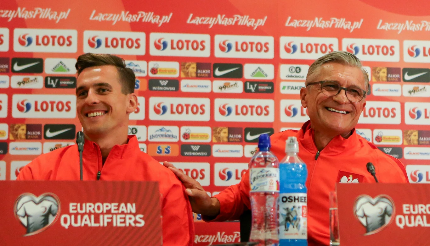 Arkadiusz Milik i Adam Nawałka. Dwóch mężczyzn ubranych w czerwone sportowe bluzy siedzi przy stole konferencyjnym, uśmiechając się szeroko, na tle ściany z logotypami sponsorów i hasłami piłkarskimi; na stole widoczne butelki wody oraz mikrofony z logo eliminacji europejskich.