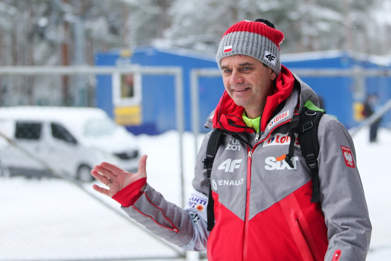 Stefan Horngacher Stefan Horngacher w czerwono-szarej kurtce i czapce z logotypami sponsorów na śniegu, z plecakiem na ramionach.