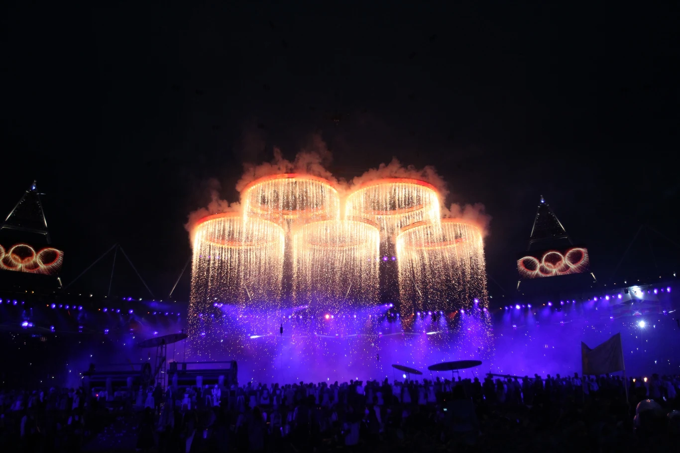 Ceremonia otwarcia - Londyn 2012 Podświetlone pierścienie olimpijskie zawieszone nad stadionem, z których sypią się iskry, otoczone fioletowym światłem i tłumem zgromadzonym poniżej.
