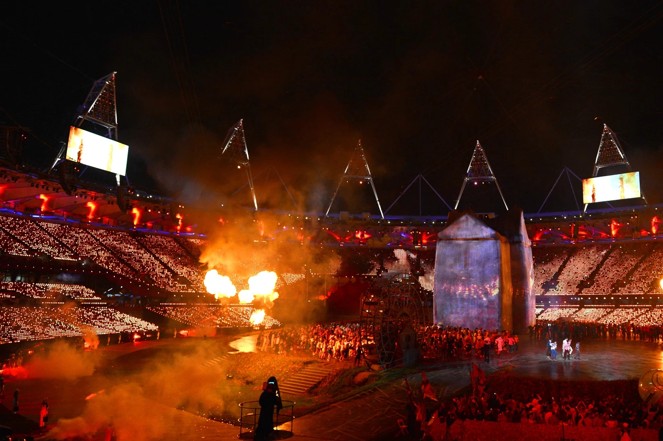 Ceremonia otwarcia - Londyn 2012 Kolorowe światła i efekty pirotechniczne rozświetlają stadion pełen widzów, centralnie uwagę przyciąga duża instalacja scenograficzna oraz tłum uczestników zgromadzonych wokół niej.