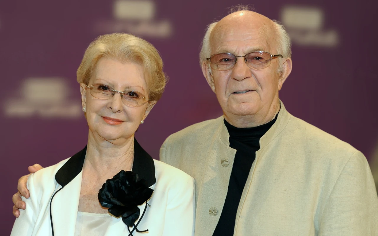 Starsza para ubrana elegancko, kobieta w białej marynarce z czarną ozdobą przy kołnierzu i okulary, mężczyzna w jasnej marynarce oraz okularach, oboje uśmiechnięci, stojący obok siebie na fioletowym tle.