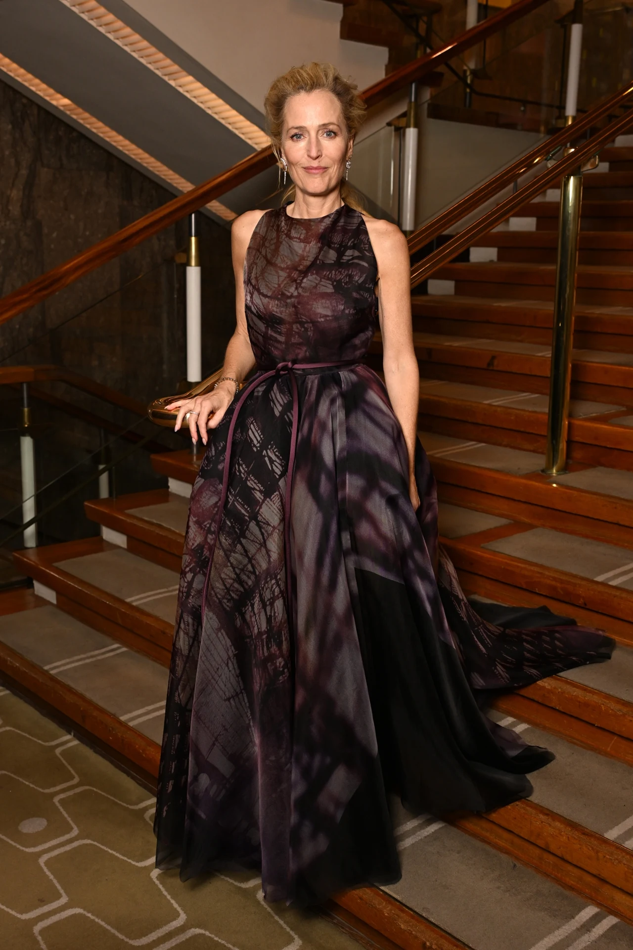 Gillian Anderson nie zniknęła po zakończeniu pracy na planie serialu. Wręcz przeciwnie, jej kariera rozkwitła Kobieta w eleganckiej, ciemnofioletowej sukni wieczorowej stoi na schodach w stylowym, nowoczesnym wnętrzu z drewnianymi poręczami i dywanem we wzory geometryczne.