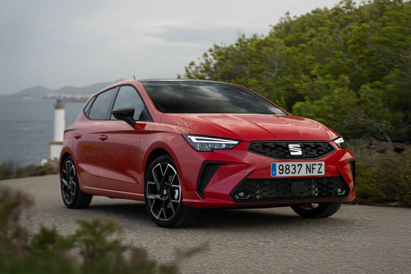 Czerwony samochód marki SEAT Ibiza, nowoczesny design, dynamiczne linie nadwozia, czarne alufelgi, zaparkowany na drodze przy zielonych krzewach.