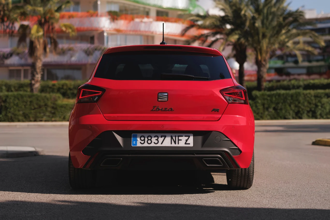 Czerwony Seat Ibiza z hiszpańską rejestracją stoi tyłem na ulicy, na tle palm i nowoczesnych budynków.
