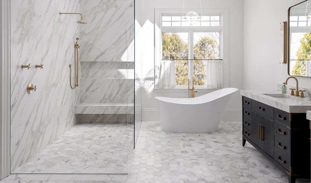 Spiek Neolith Nowoczesna łazienka z marmurowymi ścianami i podłogą, wolnostojącą wanną pod dużym oknem oraz stylową umywalką z czarną szafką i złotymi bateriami.