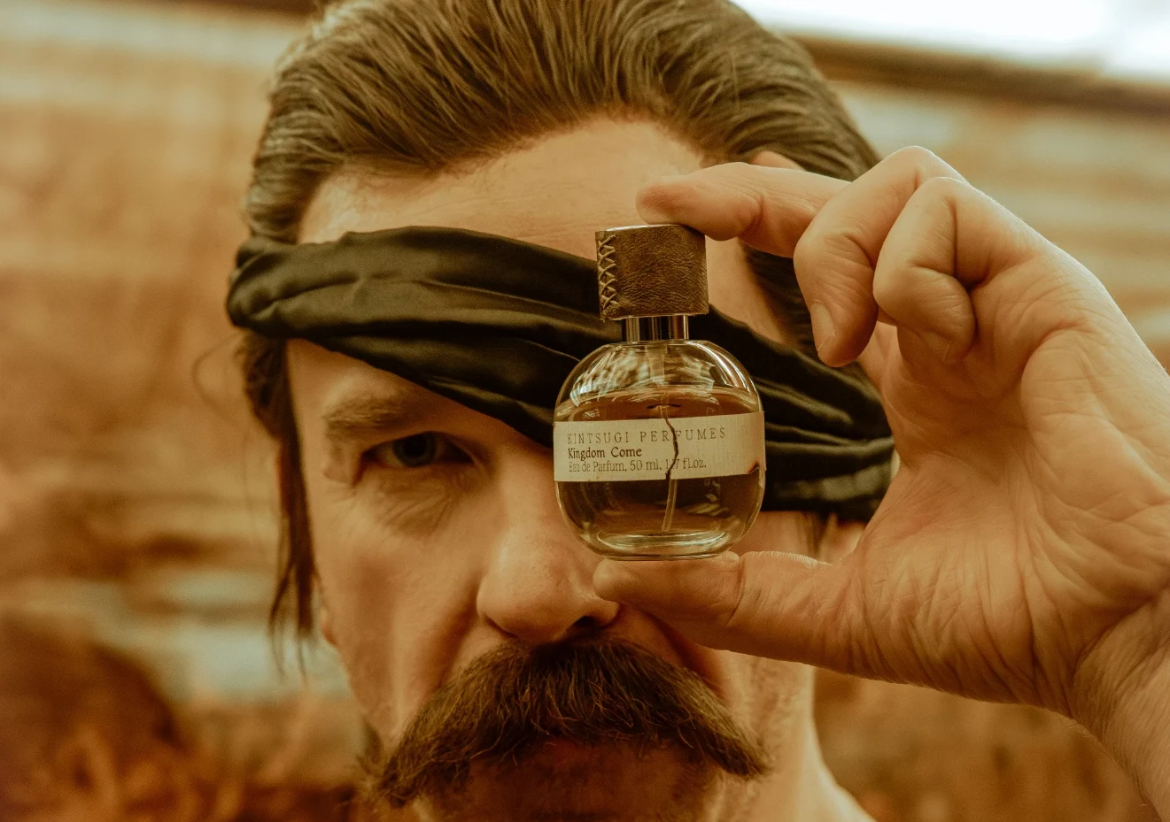 Zapach z Kingdom Come / kingdomcome-store Mężczyzna z wąsem i opaską na oczach trzyma flakon perfum Kingdom Come przed twarzą.