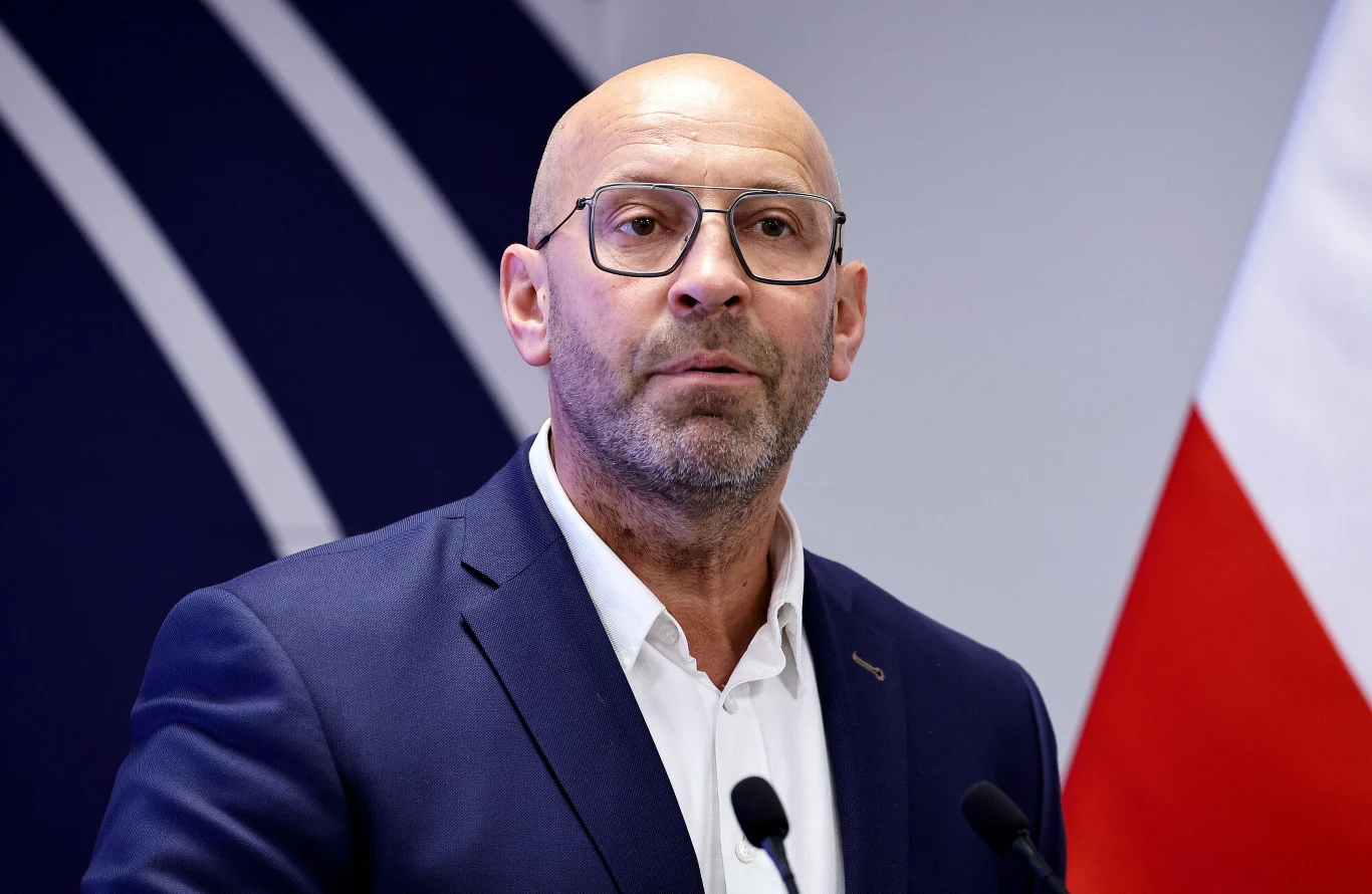 Jakub Rutnicki Jakub Rutnicki w okularach, w granatowej marynarce i białej koszuli, stoi przy mównicy na tle fragmentu flagi Polski.
