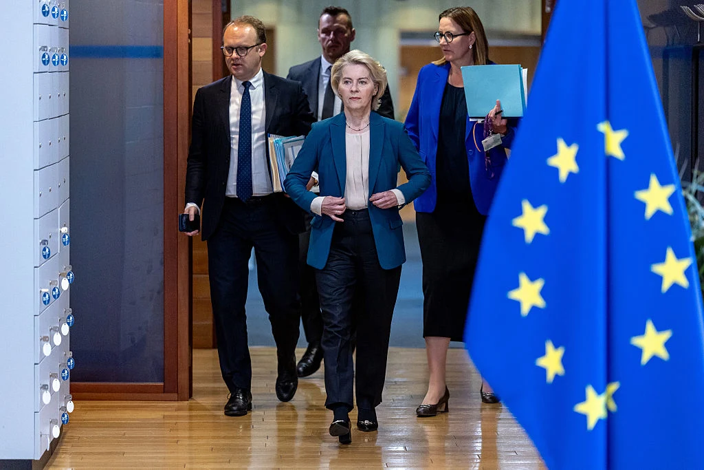 Kobieta w niebieskim żakiecie idzie zdecydowanym krokiem korytarzem w towarzystwie trzech osób ubranych biznesowo, z przodu widoczna flaga Unii Europejskiej.