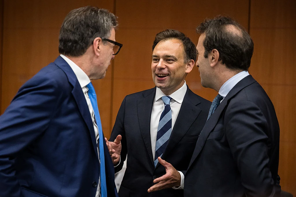 Trzech mężczyzn w garniturach prowadzi rozmowę w jasnym, biurowym wnętrzu; centralna postać uśmiecha się i gestykuluje dłonią, skupiając uwagę pozostałych rozmówców.