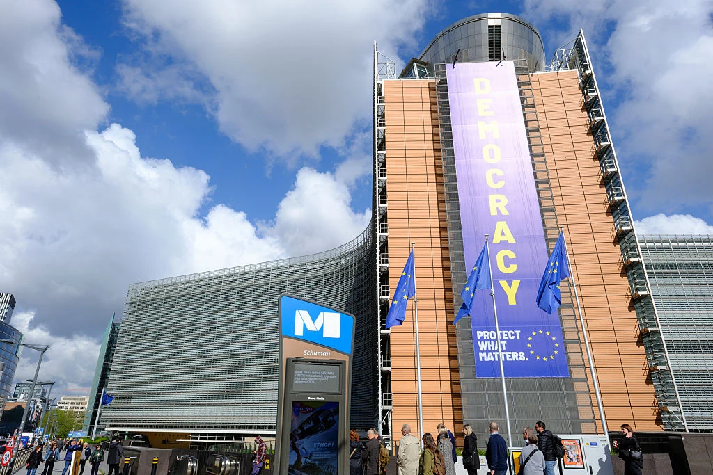 Nowoczesny budynek z wielkim żółtym transparentem z napisem 'DEMOCRACY' oraz trzema powiewającymi flagami Unii Europejskiej, przed wejściem grupka ludzi i znak stacji metra.