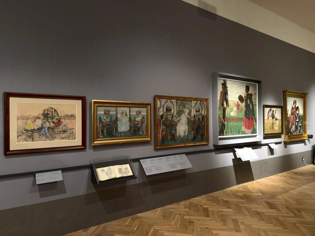 Kilka obrazów w złotych i brązowych ramach wiszących na szarej ścianie muzeum, przedstawiających postacie w tradycyjnych strojach oraz sceny wiejskie, poniżej ekspozycja szkiców w gablotach, drewniana podłoga parkietowa.