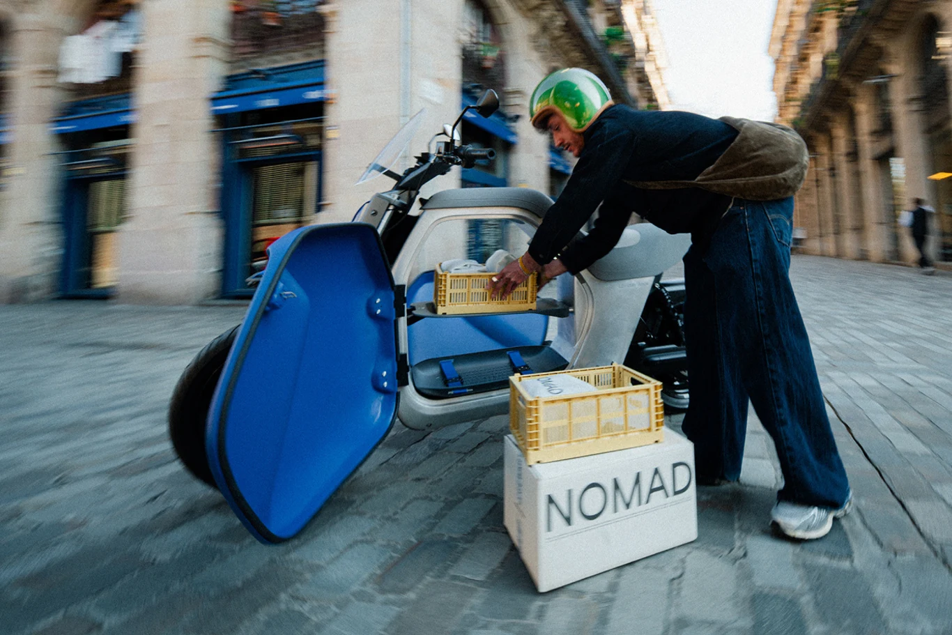 Mężczyzna w kasku wyjmuje plastikowe skrzynki z bagażnika nietypowego skutera zaparkowanego na brukowanej ulicy, obok znajduje się karton z napisem NOMAD.