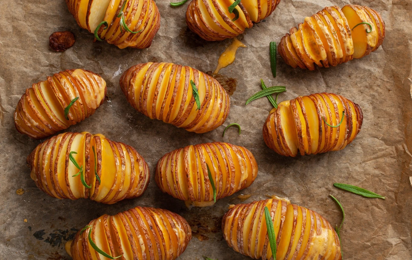 Ziemniaki Hasselback mogą być zjadane solo albo z dodatkami Plastry ziemniaków pieczone metodą Hasselback z kawałkami sera oraz gałązkami świeżego rozmarynu, ułożone na arkuszu papieru do pieczenia.