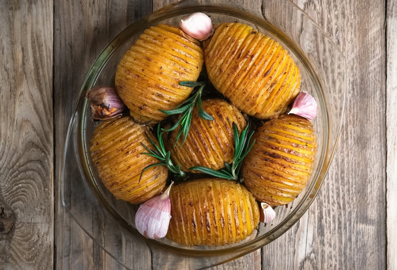 Ziemniaki Hasselback trzeba piec około godziny Złociste, pieczone ziemniaki hasselback udekorowane świeżym rozmarynem i ząbkami czosnku w szklanym naczyniu na drewnianym stole.