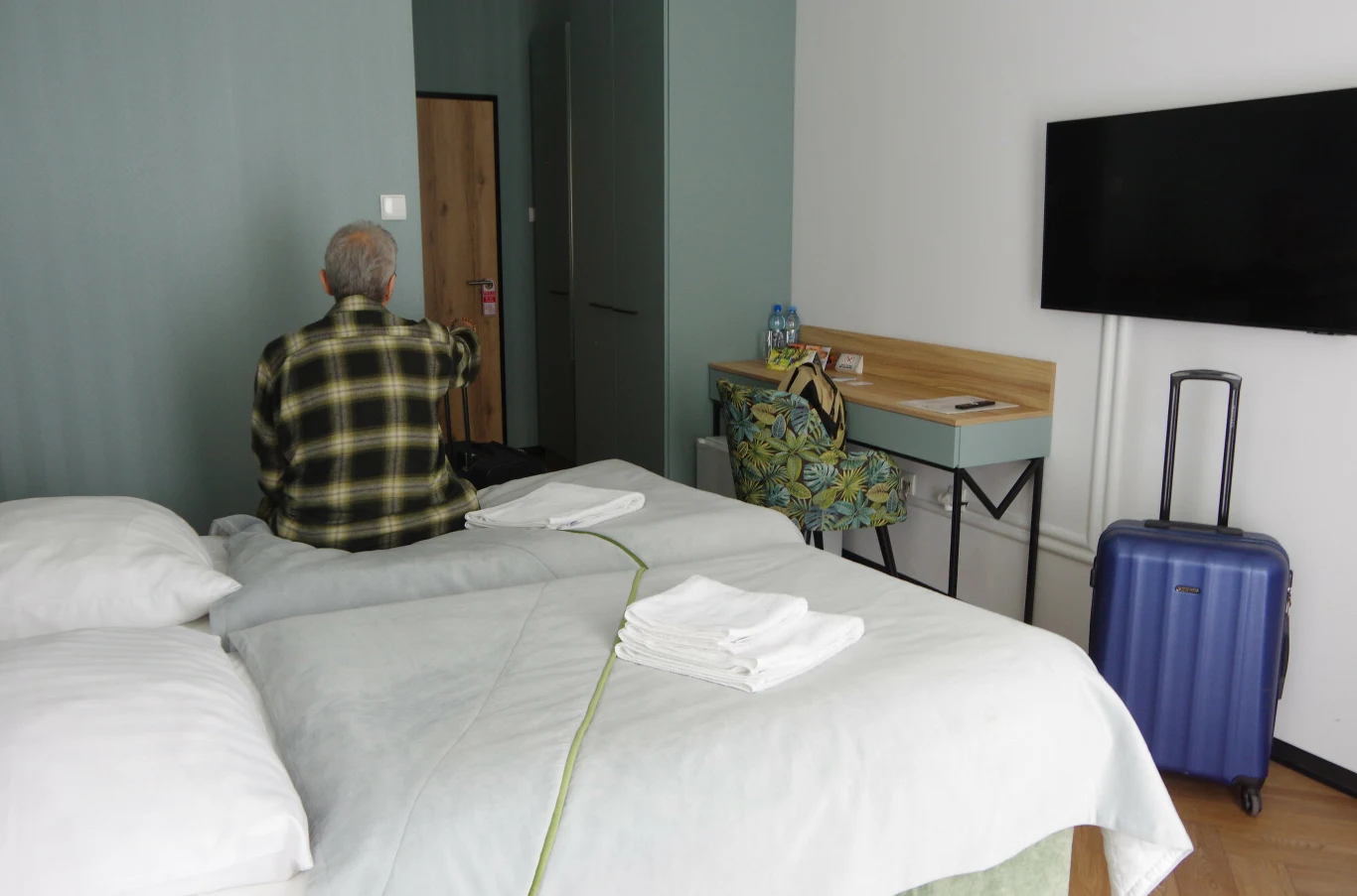 "Jedynek" w sanatoriach jest niewiele. Starszy mężczyzna w koszuli w kratę siedzi na łóżku w pokoju sanatorium, obok leżą białe ręczniki i walizka.