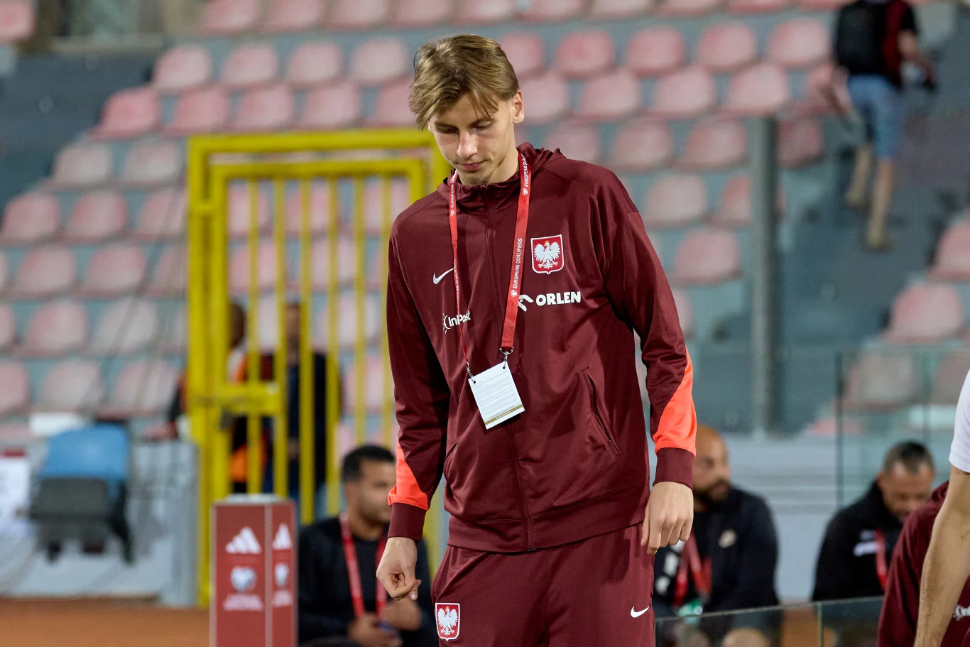 Młody mężczyzna w bordowym dresie reprezentacji Polski z identyfikatorem na szyi stoi na stadionie sportowym, za nim widać kilka osób w podobnych strojach.