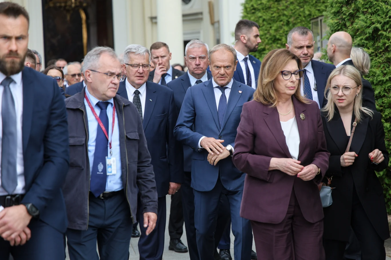 Grupa elegancko ubranych osób idących razem podczas oficjalnego wydarzenia na zewnątrz, wśród nich kobieta w bordowym garniturze i mężczyzna w niebieskim garniturze, otoczeni przez innych uczestników oraz ochronę.