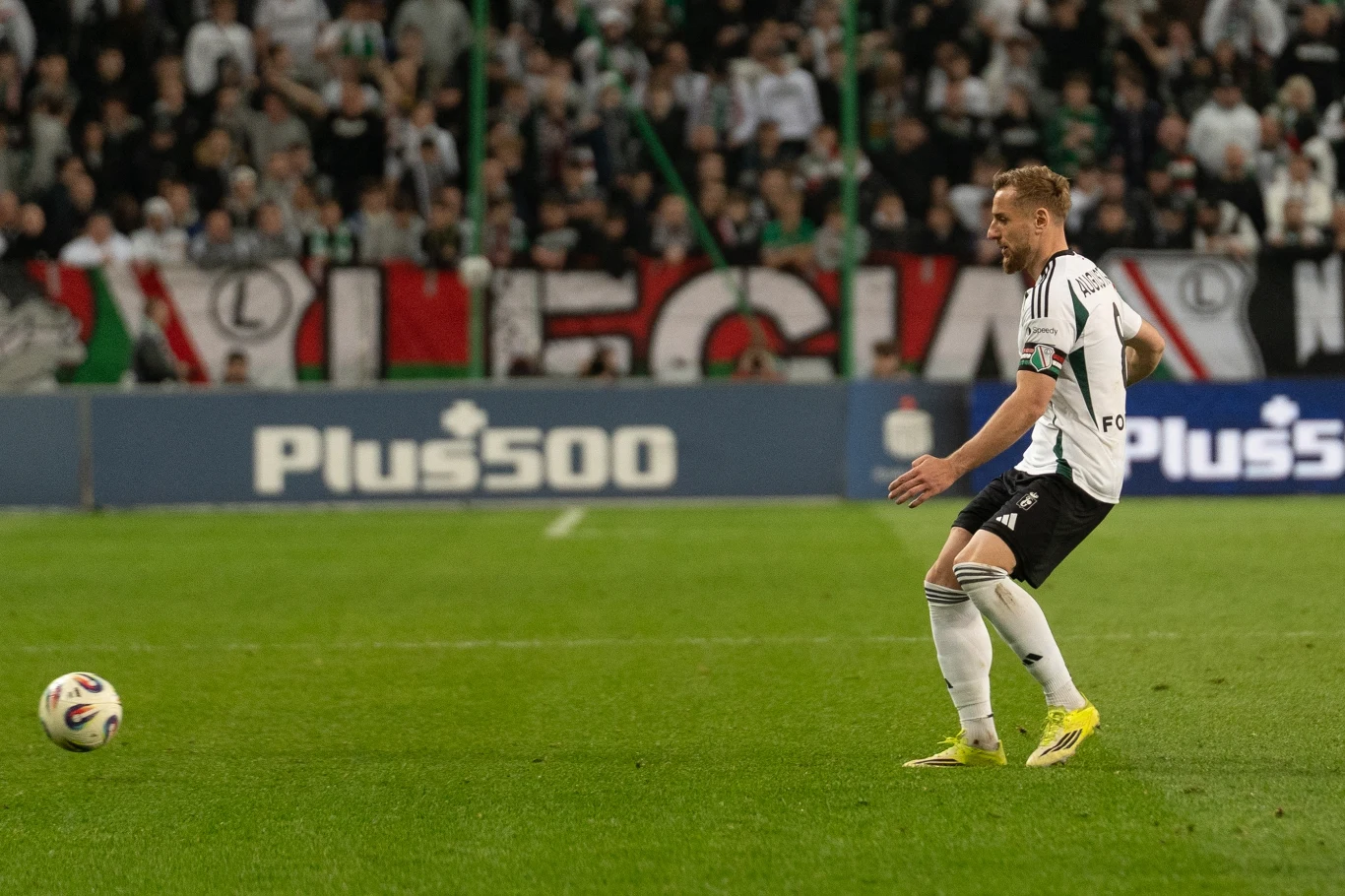 Piłkarz w białym stroju z logiem Legii Warszawa kontroluje piłkę podczas meczu na stadionie, w tle trybuny pełne kibiców z widocznym transparentem 'LEGIA'.
