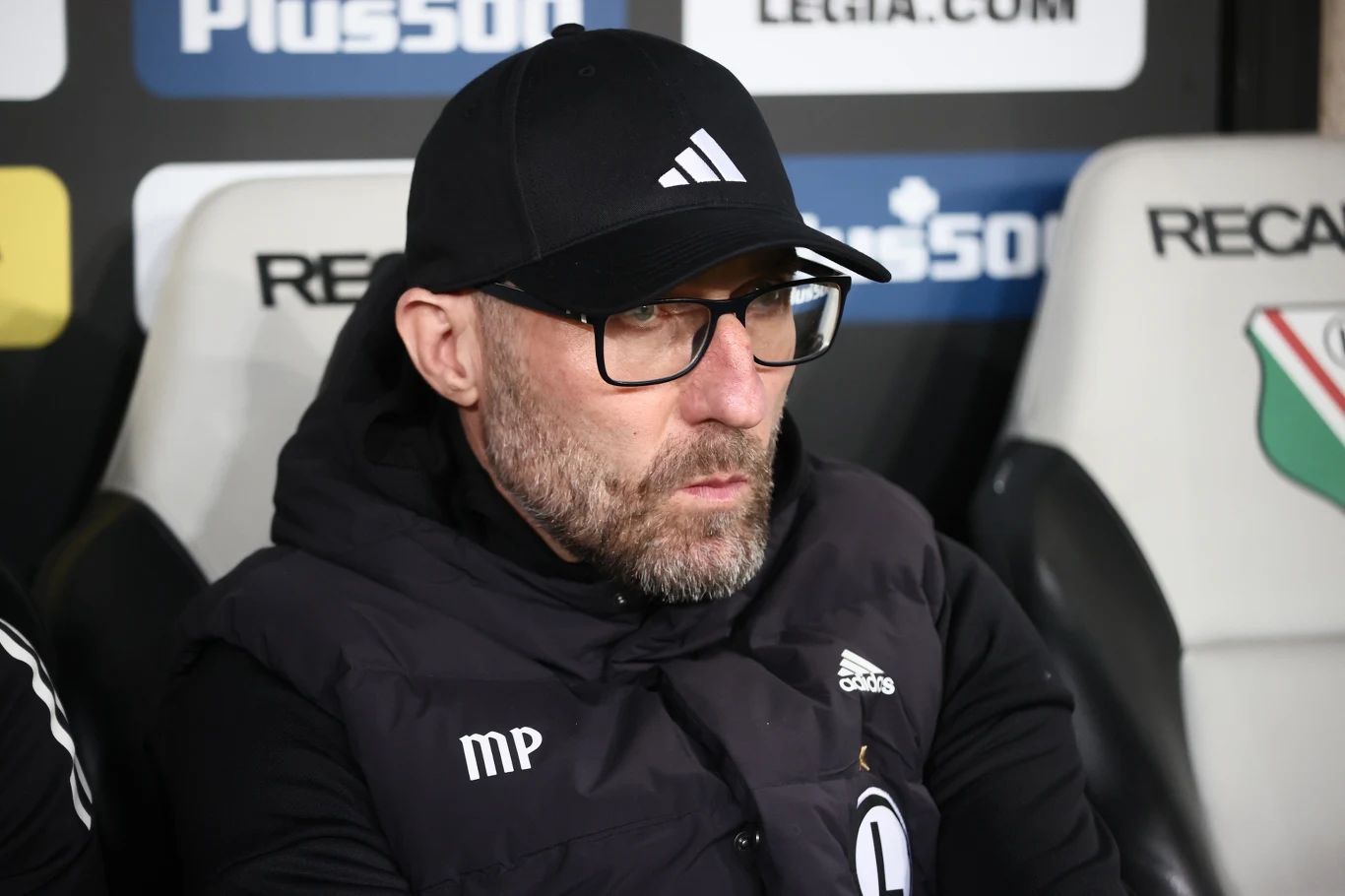 Mężczyzna w czarnej czapce z daszkiem i okularach, ubrany w sportową bluzę z logo znanej marki, siedzi na ławce rezerwowych obok oznaczenia klubu piłkarskiego Legia Warszawa.