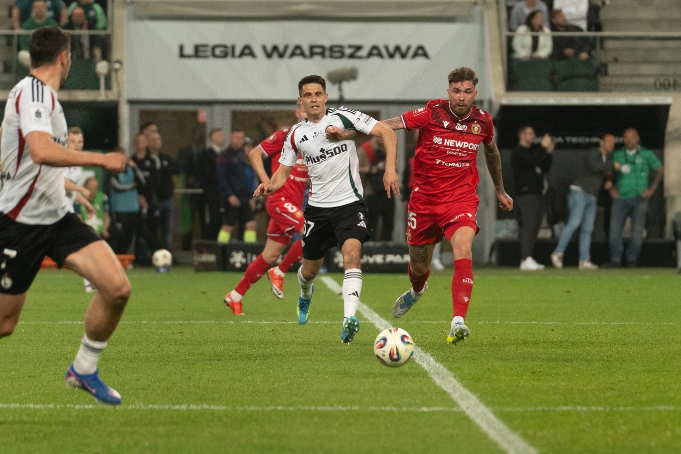 Dwóch piłkarzy walczy o piłkę na murawie stadionu Legii Warszawa, w tle widoczna ławka rezerwowych oraz kibice na trybunach.