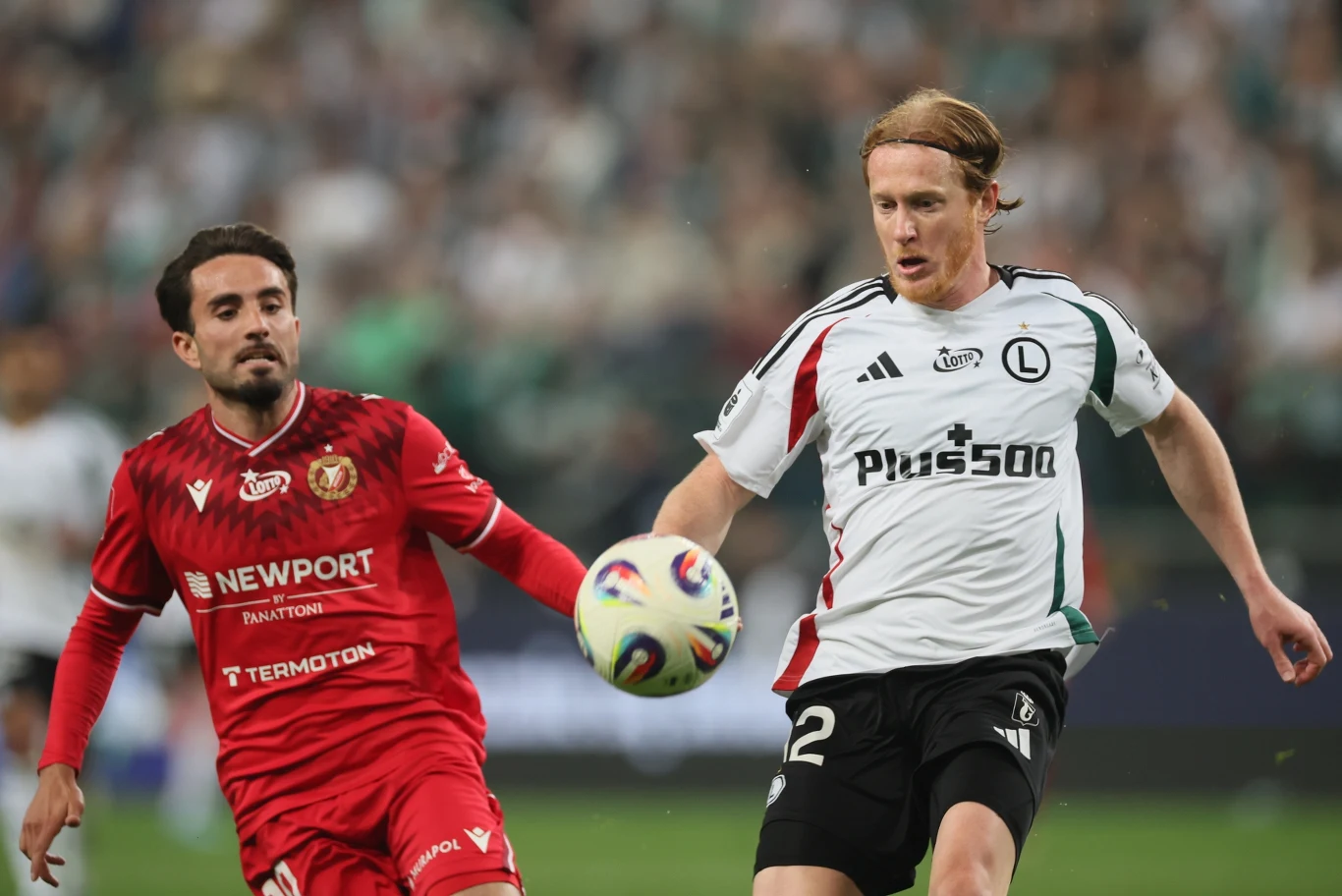 Dwóch piłkarzy w dynamicznej walce o piłkę, zawodnik w białej koszulce Legii Warszawa prowadzi futbolówkę, podczas gdy gracz w czerwonym stroju rywala próbuje odebrać mu piłkę.