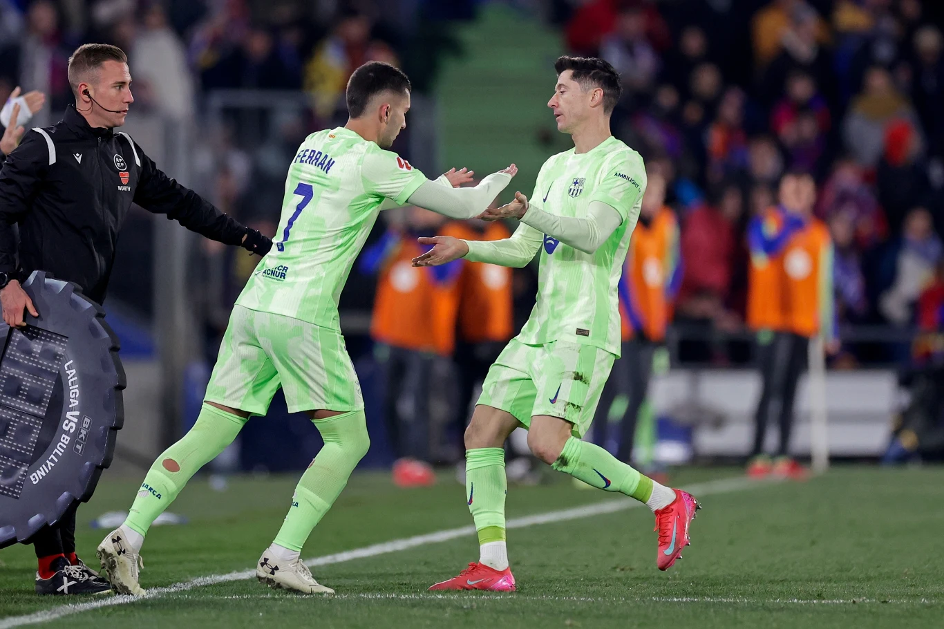 Ferran Torres i Robert Lewandowski Dwóch piłkarzy w jasnozielonych strojach wykonuje zmianę na boisku podczas meczu piłki nożnej, obok stoi sędzia techniczny z tablicą do zmian, w tle widoczni są kibice i członkowie obsługi boiska.