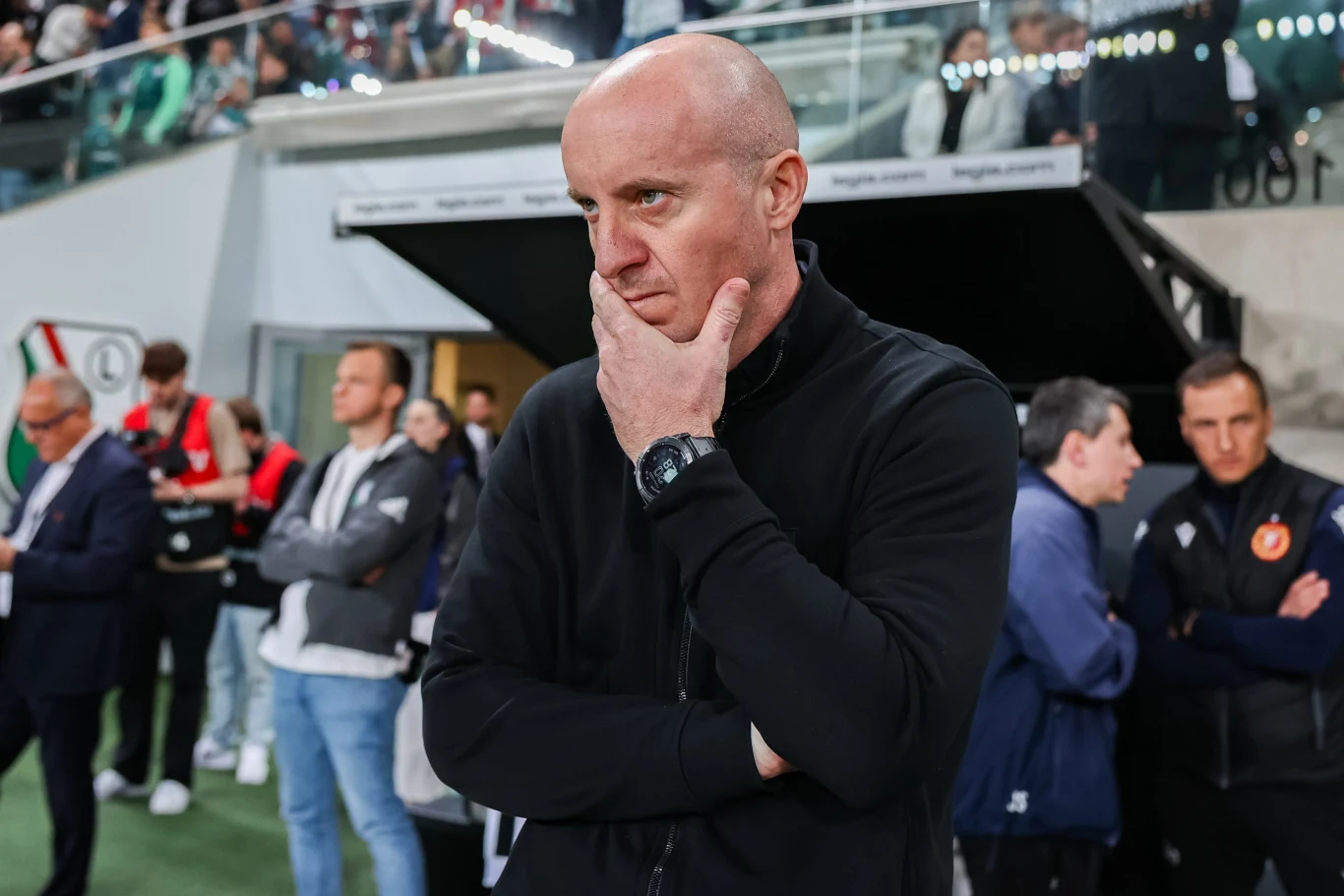 Aleksandar Vuković Łysy mężczyzna w ciemnej bluzie stoi zamyślony z ręką opartą na brodzie, na tle stadionu i skupionych osób w sportowych strojach.
