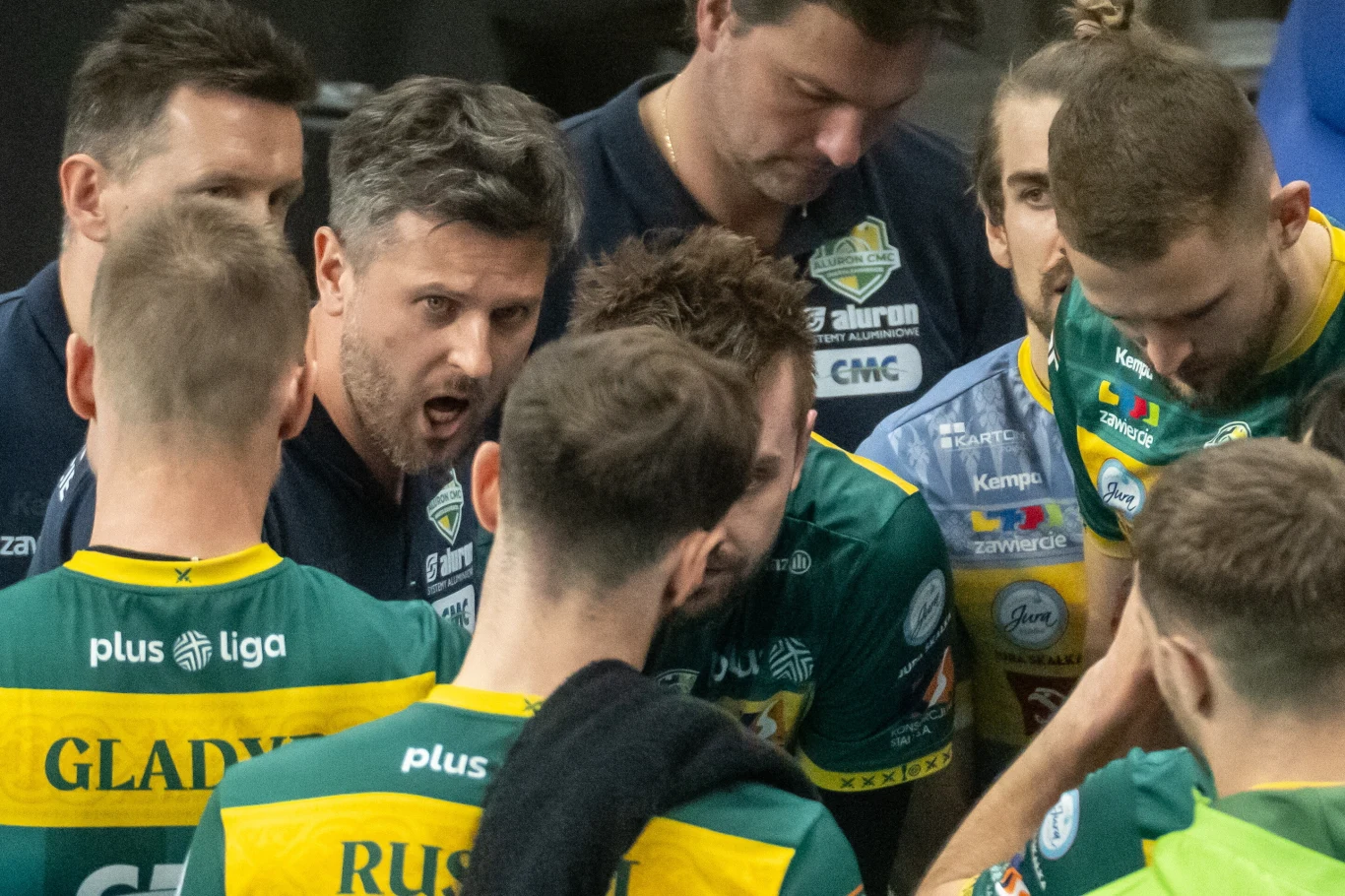 Trener drużyny siatkarskiej motywuje zawodników podczas przerwy, otoczony przez graczy w zielono-żółtych strojach z widocznymi nazwiskami na plecach.