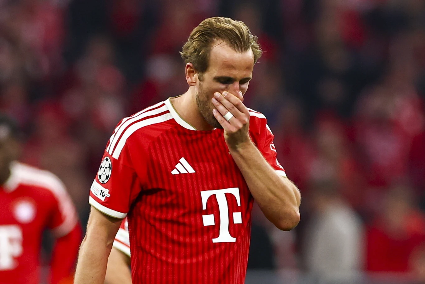 Piłkarz Bayernu Monachium w czerwonej koszulce z widocznym logo sponsora zasłania twarz dłonią w geście rozczarowania na tle rozmytego stadionu.