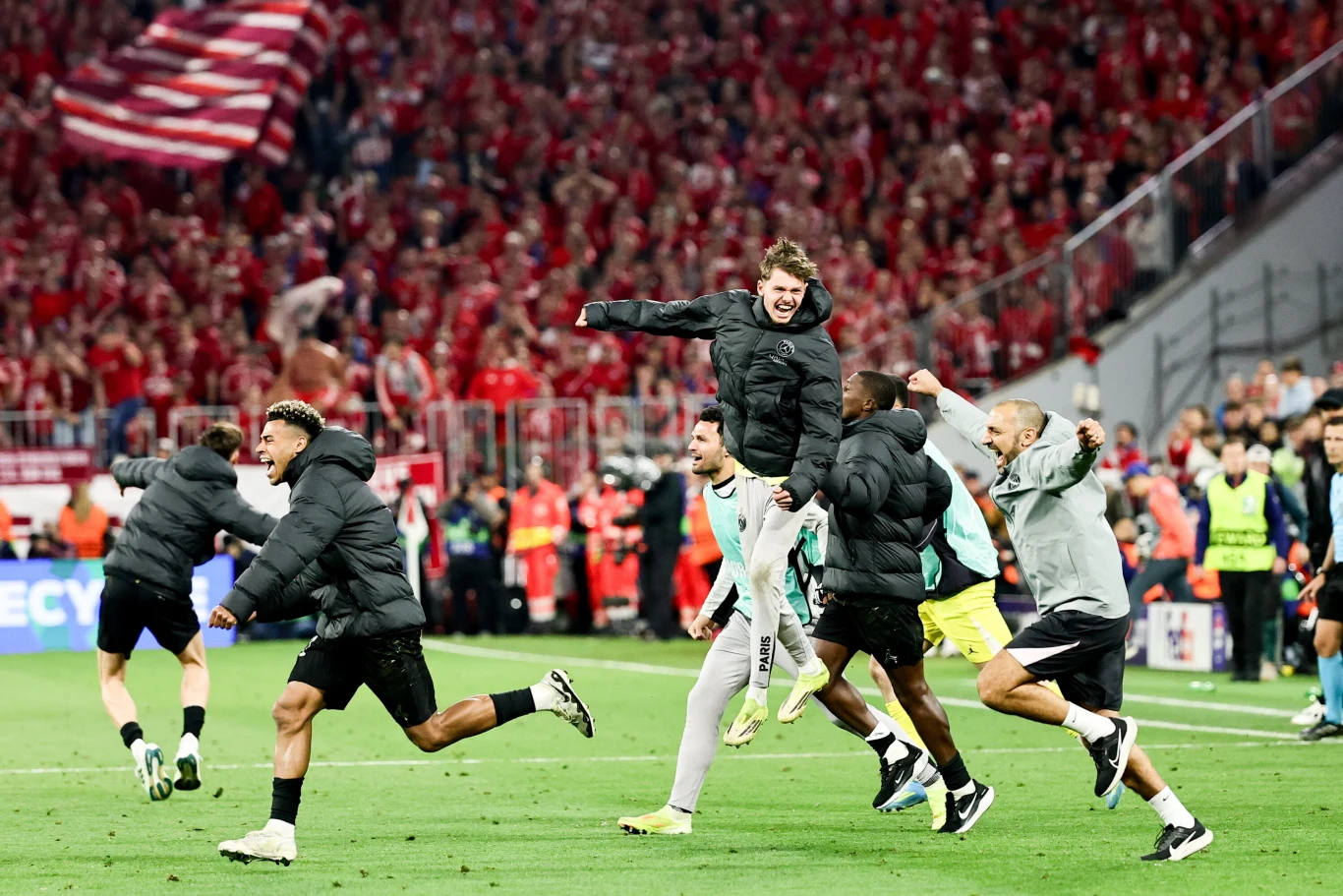 Grupa piłkarzy biegnących z uniesionymi rękami po murawie stadionu podczas świętowania wśród tłumu kibiców ubranych na czerwono.