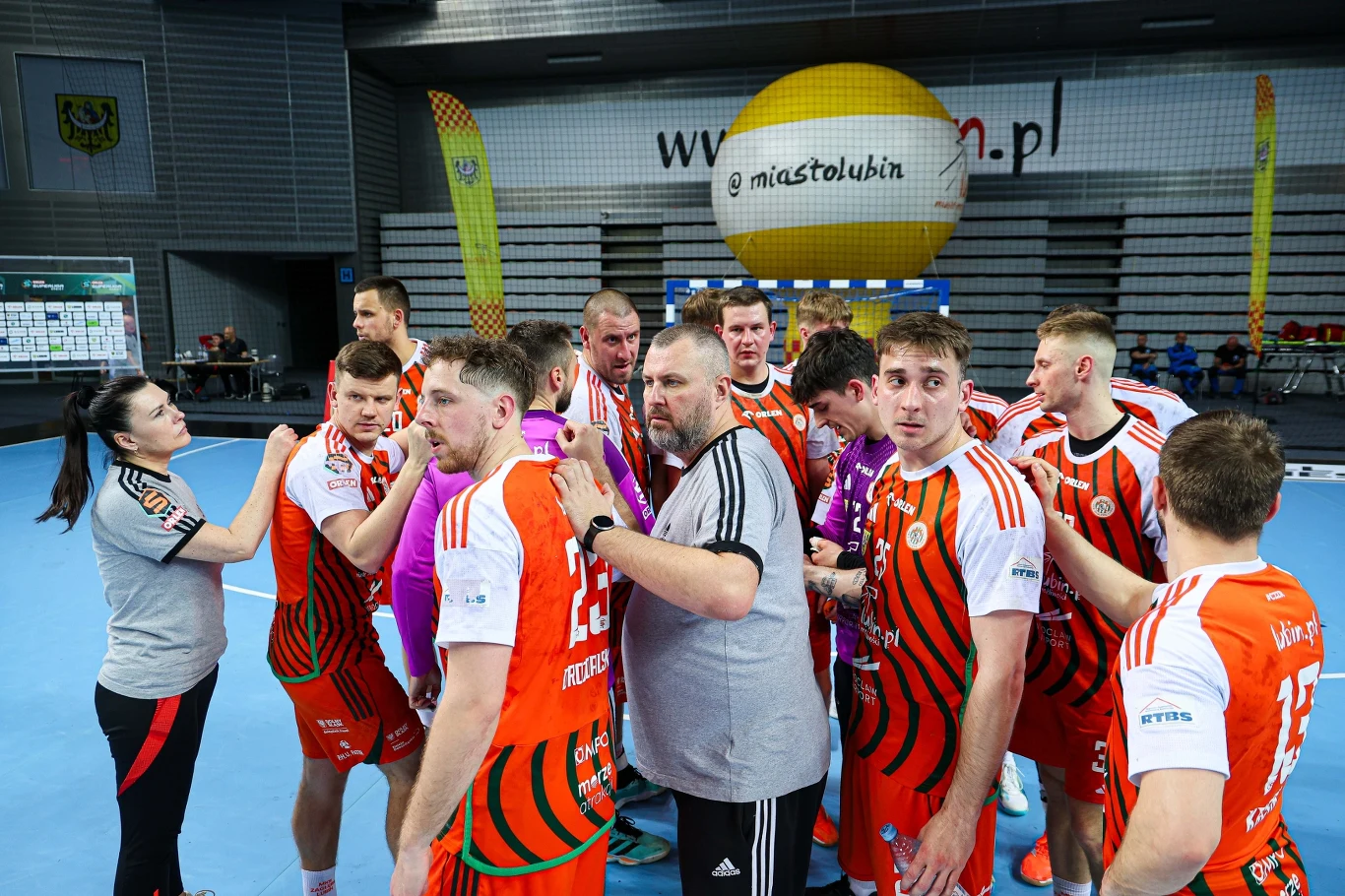 MKS Zagłębie Lubin Drużyna piłki ręcznej w pomarańczowych strojach zebrana w kręgu podczas przerwy na hali sportowej, trener przekazuje uwagi zawodnikom.