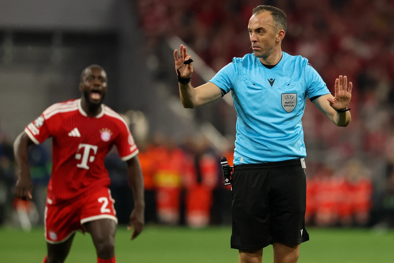 Bayern - PSG Sędzia piłkarski w niebieskim stroju sygnalizuje decyzję, obok piłkarz Bayernu Monachium w czerwonym stroju.