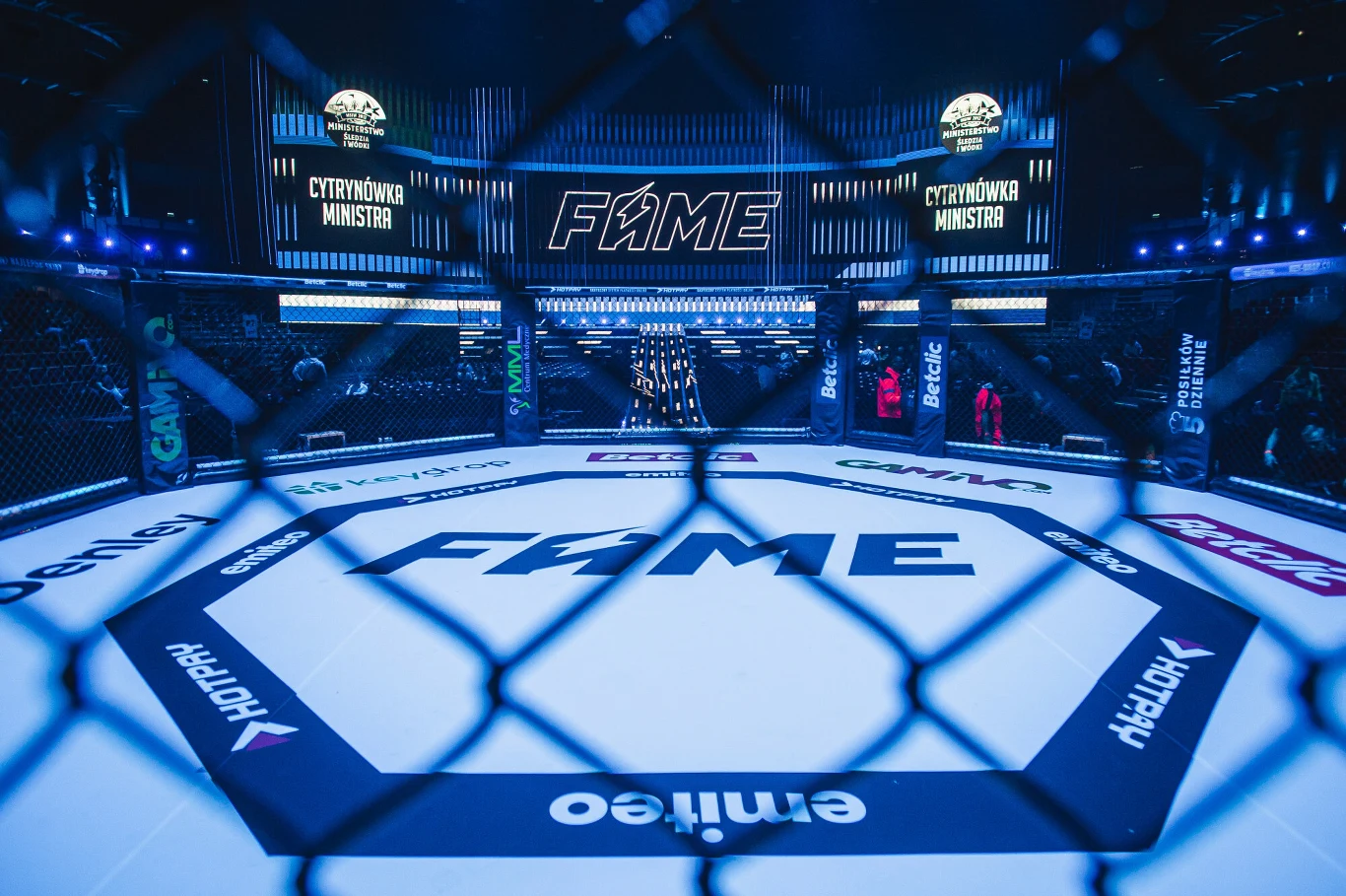 Oktagon podczas jedne z gal FAME MMA Oktagonalna klatka federacji Fame MMA oświetlona niebieskimi światłami, na środku widoczne logo organizacji, w tle telebimy i elementy scenografii galowej.