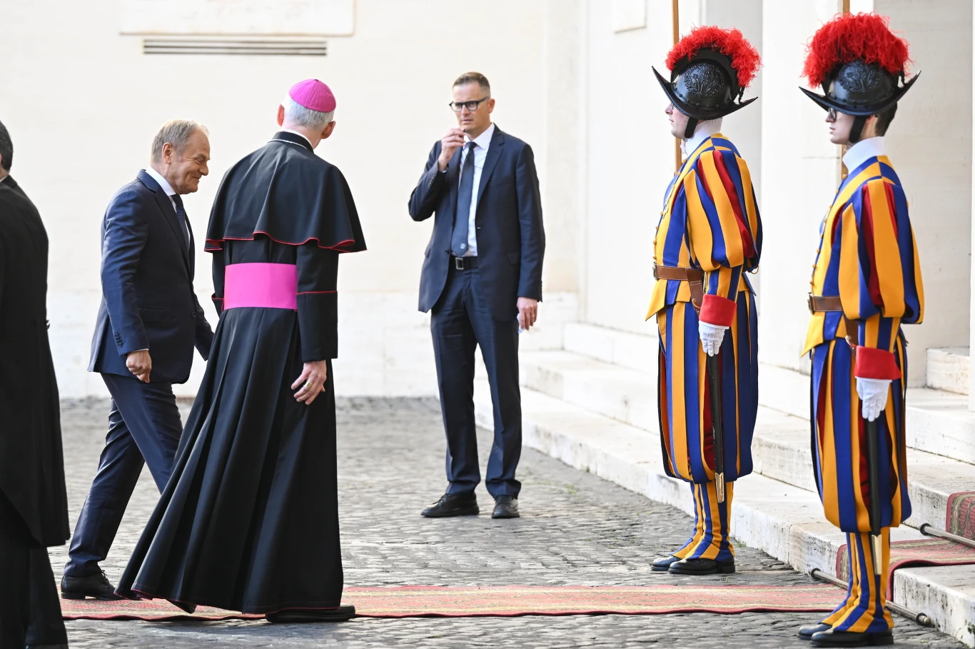 Premier Donald Tusk i duchowny katolicki zbliżają się do szwajcarskich gwardzistów przed Watykanem.