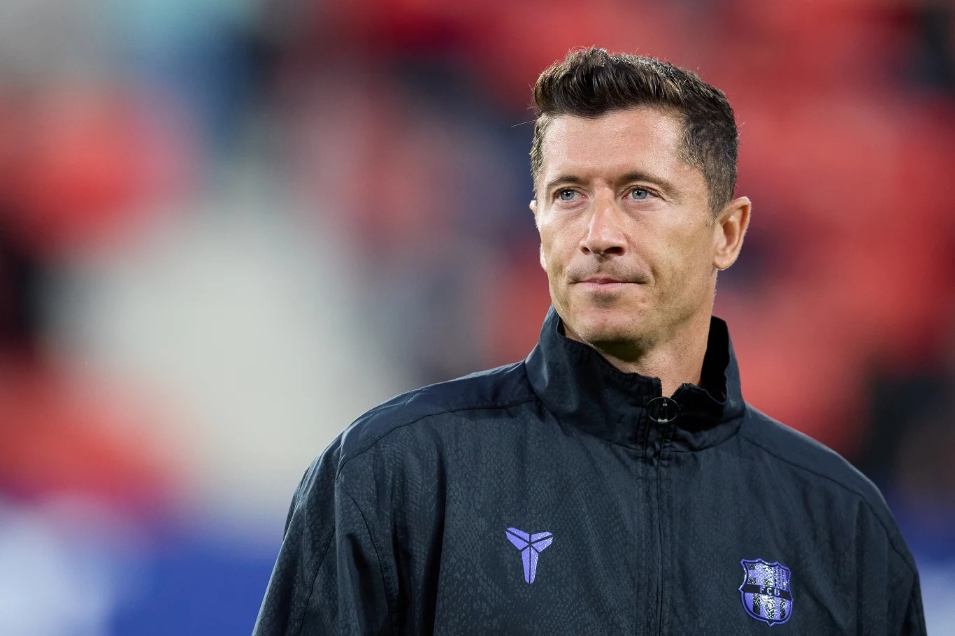 Robert Lewandowski Robert Lewandowski w sportowej kurtce z herbem drużyny na tle rozmytego, czerwonego stadionu.
