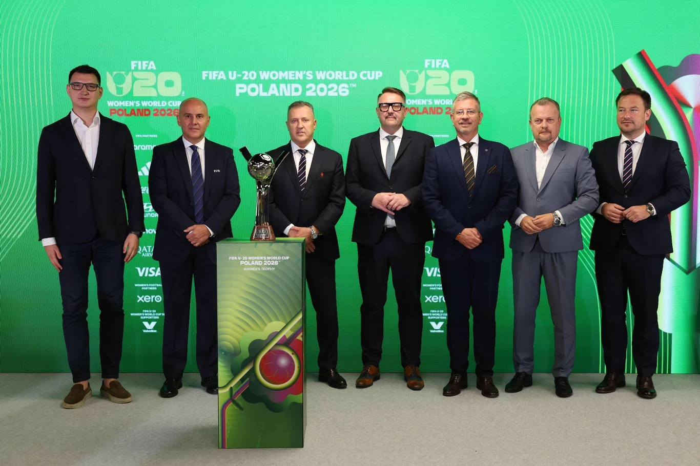 Siedmiu mężczyzn w garniturach i eleganckich ubraniach pozuje obok trofeum mistrzostw świata FIFA U-20 kobiet, stojąc na tle zielonej ścianki z logo turnieju i napisem 'FIFA U-20 Women’s World Cup Poland 2026'.