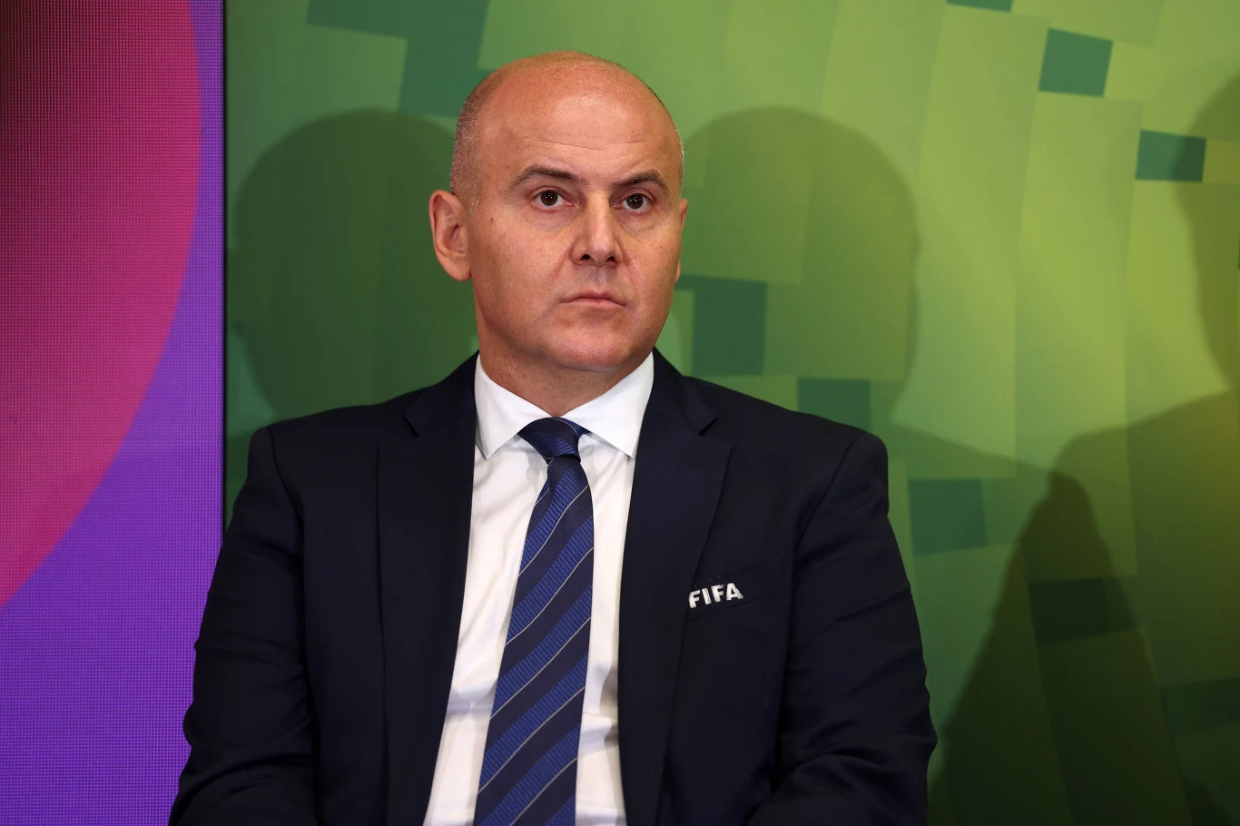 Łysy mężczyzna ubrany w ciemny garnitur z krawatem i białą koszulą, z logo FIFA na marynarce, siedzi na tle kolorowej, geometrycznej grafiki.