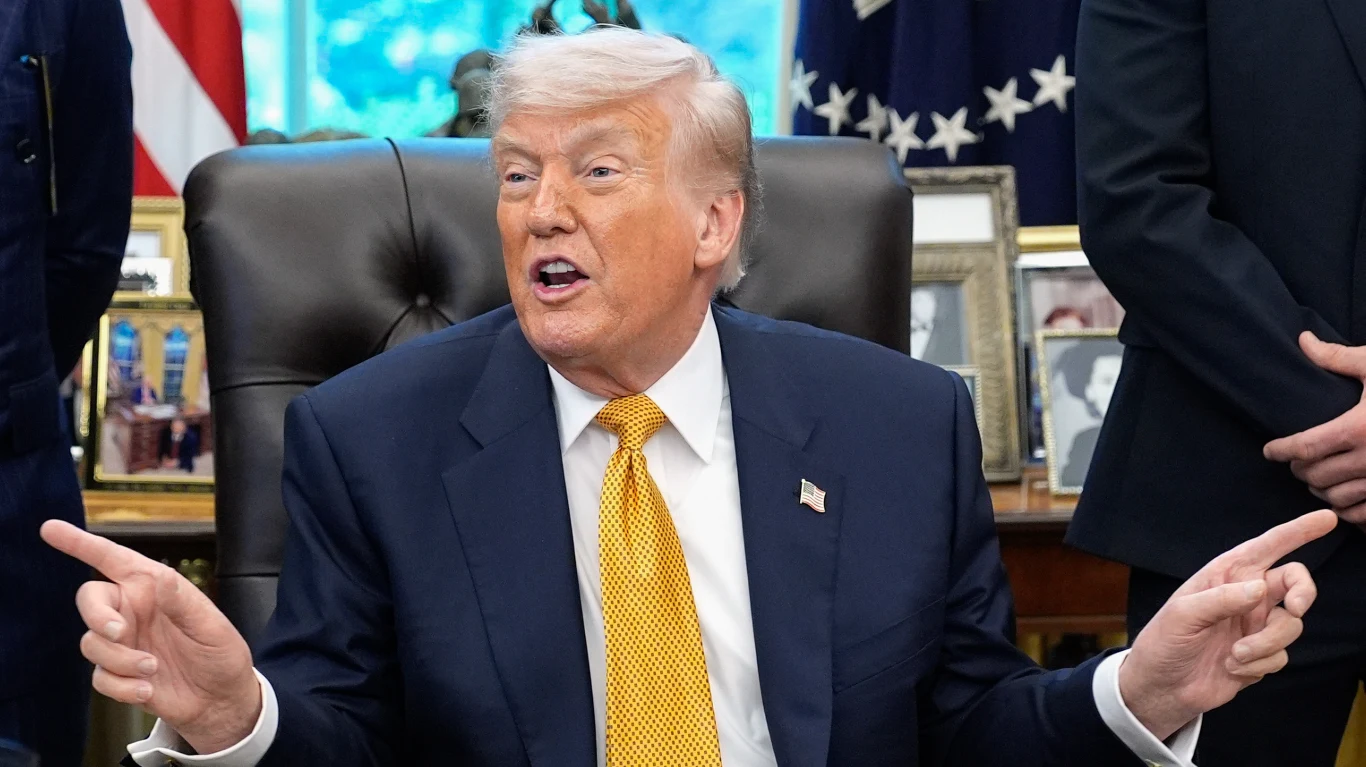 Donald Trump Donald Trump w garniturze i żółtym krawacie przy biurku, gestykuluje, w tle flagi i zdjęcia w ramkach.