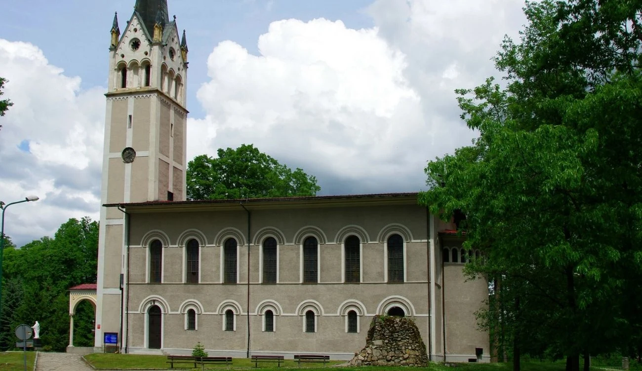 Kościół z wysoką wieżą zwieńczoną iglicą, ozdobiony rzędem półkolistych okien, otoczony zielenią drzew i krzewów, na pierwszym planie niewielki kamienny kopiec.