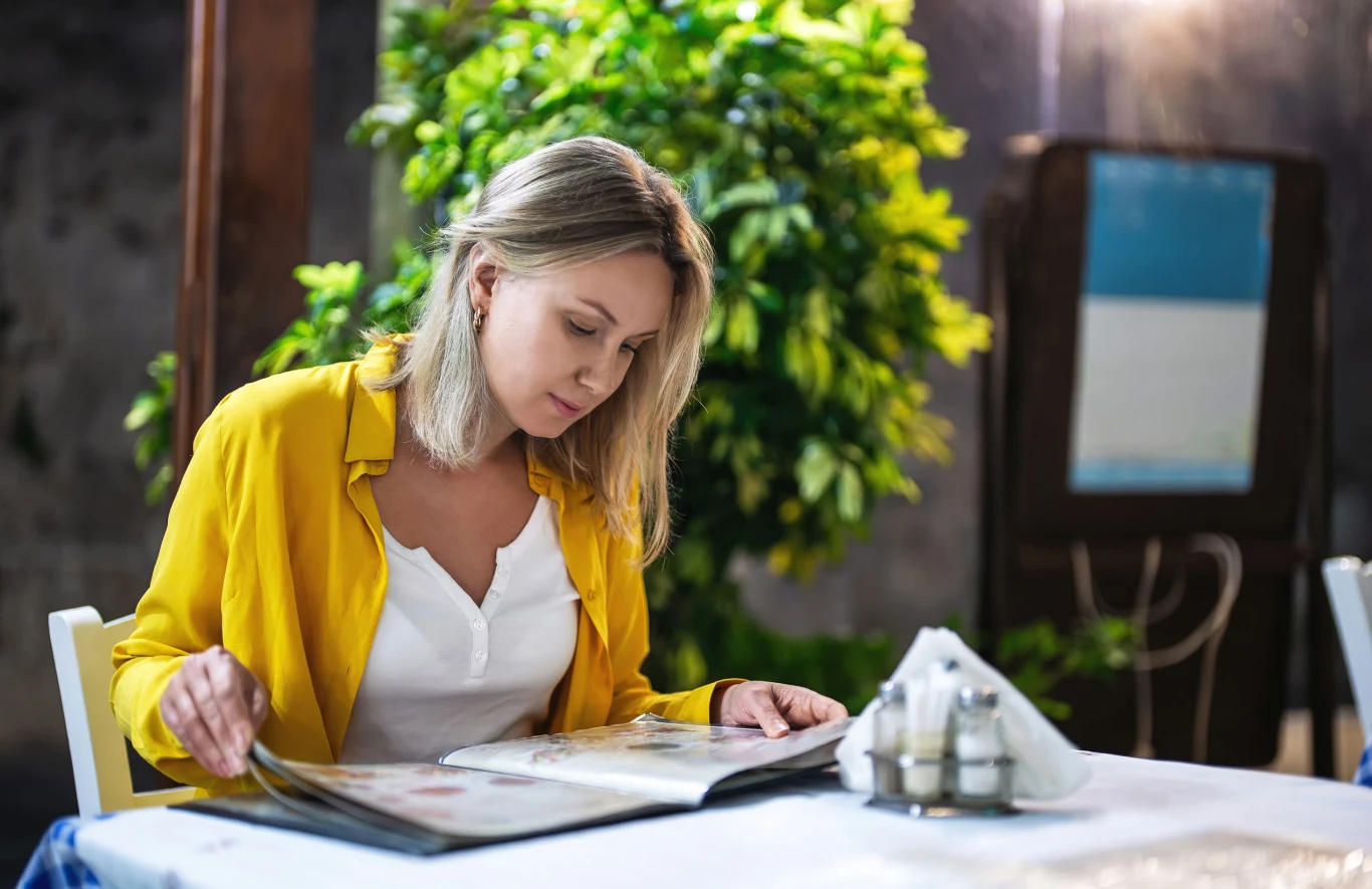 Uważaj, z kim idziesz na kolację. Te znaki zodiaku potrafią narobić wstydu Kobieta w żółtej koszuli siedzi przy stole w restauracji i przegląda menu; na stole serwetki i przyprawy.
