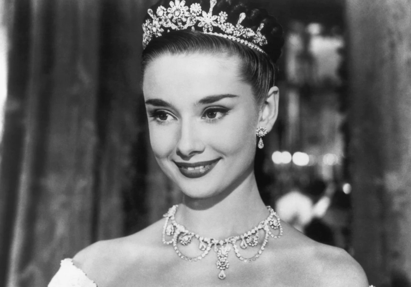 Jak pachniały perfumy Audrey Hepburn Młoda kobieta w diamentowej tiarze, balowej sukni z odkrytymi ramionami i eleganckiej biżuterii.