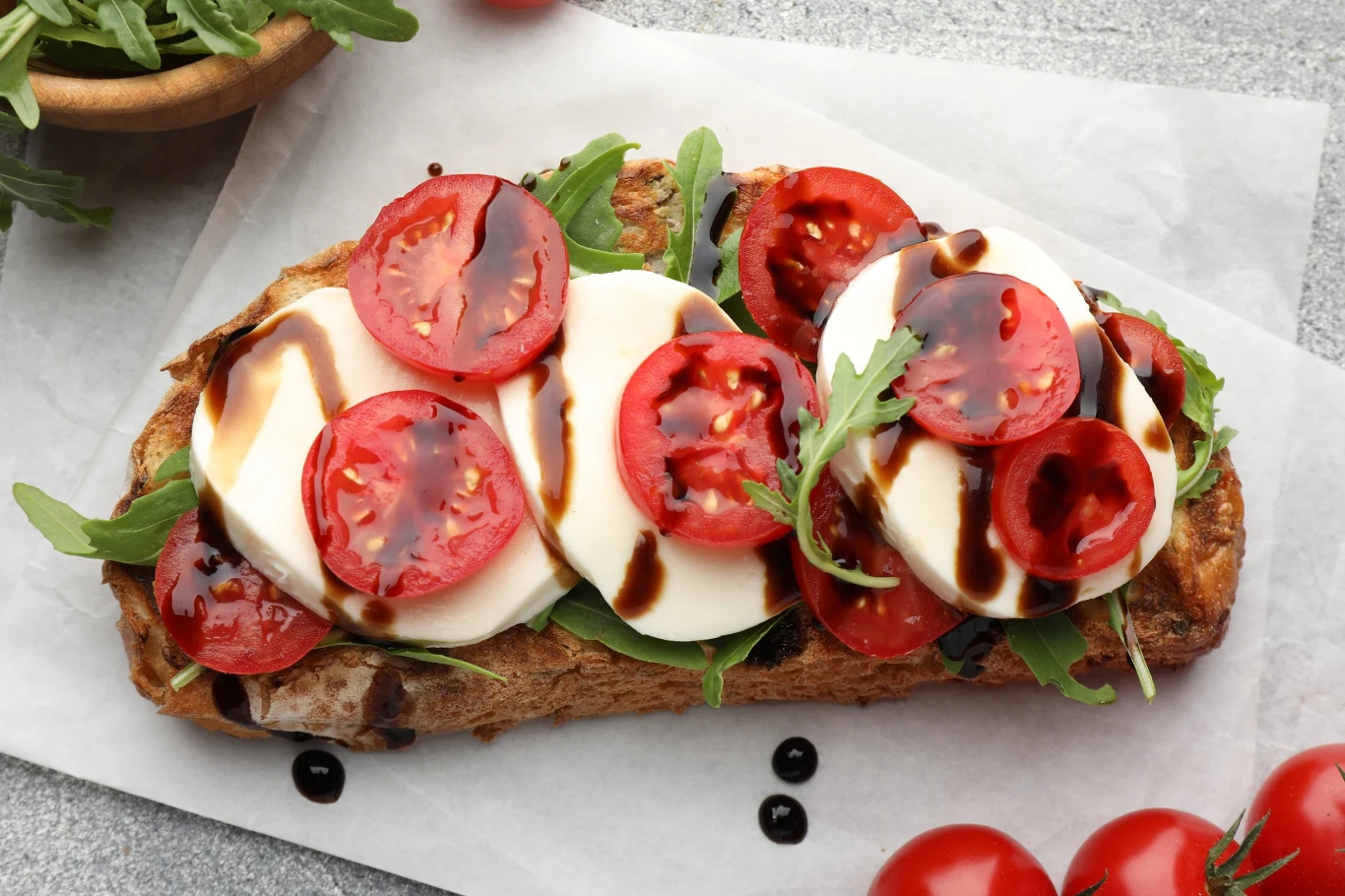 Grzanka z chrupiącego pieczywa z plastrami mozzarelli, świeżymi pomidorami, rukolą i sosem balsamicznym.
