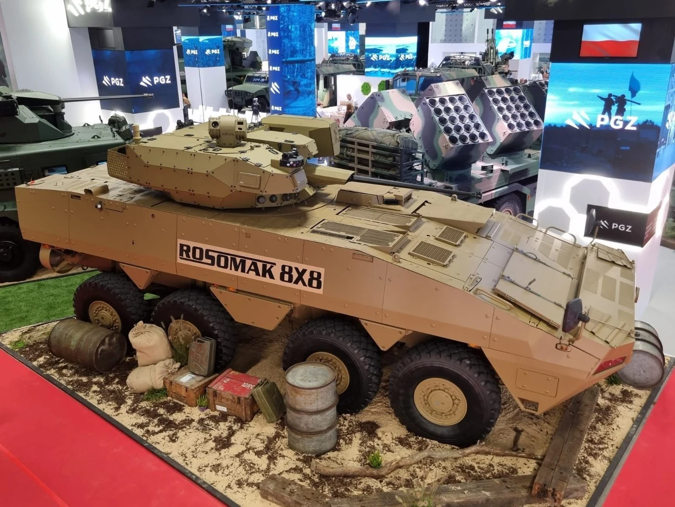 Rosomak-L ze słowacką wieżą Turra 30 Pojazd opancerzony Rosomak 8x8 w piaskowym kamuflażu ustawiony na pokazowym stanowisku otoczonym workami z piaskiem, beczkami i drewnianymi belkami, w tle ekspozycja innych pojazdów wojskowych oraz podświetlane panele z logo PGZ.