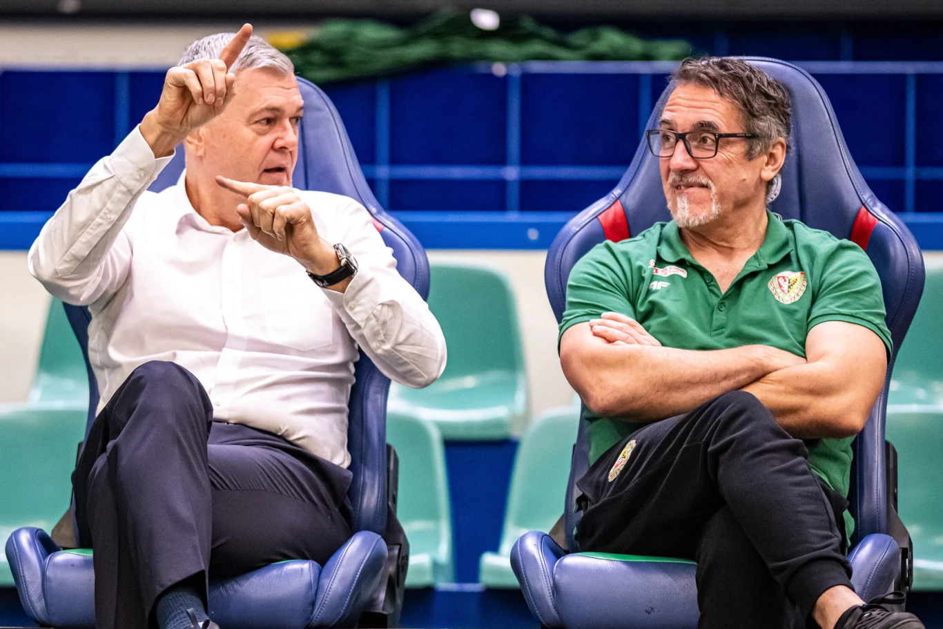 Dwóch mężczyzn siedzi obok siebie na stadionowych fotelach, jeden w białej koszuli gestykuluje, drugi w zielonej koszulce z logo sportowego klubu obserwuje go z zainteresowaniem.