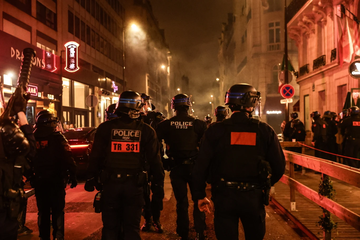 Grupa policjantów w pełnym umundurowaniu taktycznym stoi na oświetlonej ulicy miasta, w tle widoczne są budynki, światła i unoszący się dym.
