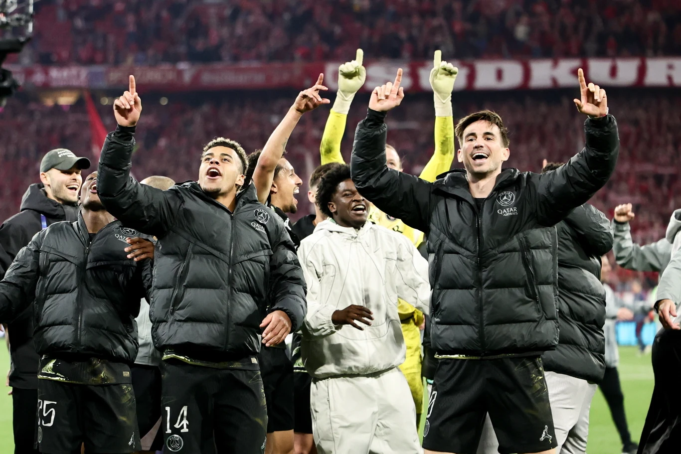 Grupa piłkarzy w czarnych kurtkach świętuje na boisku z uniesionymi rękami, wyrażając radość i triumf po meczu, w tle tłum kibiców i stadionowe światła.