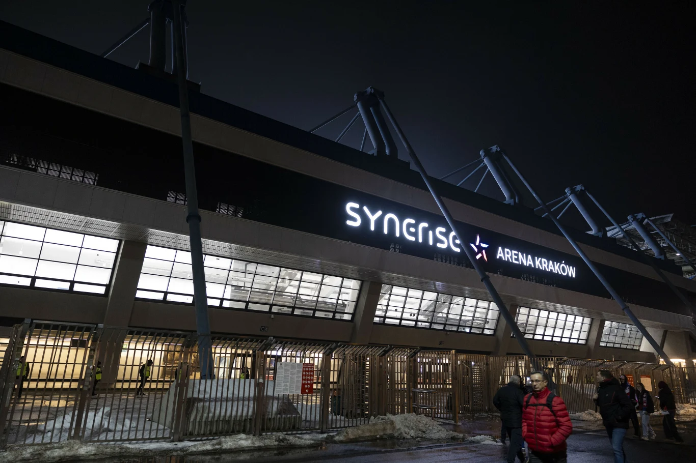 Synerise Arena Kraków Podświetlony napis Synerise Arena Kraków na elewacji nowoczesnego budynku, przed którym przechodzą osoby ubrane w zimowe kurtki.